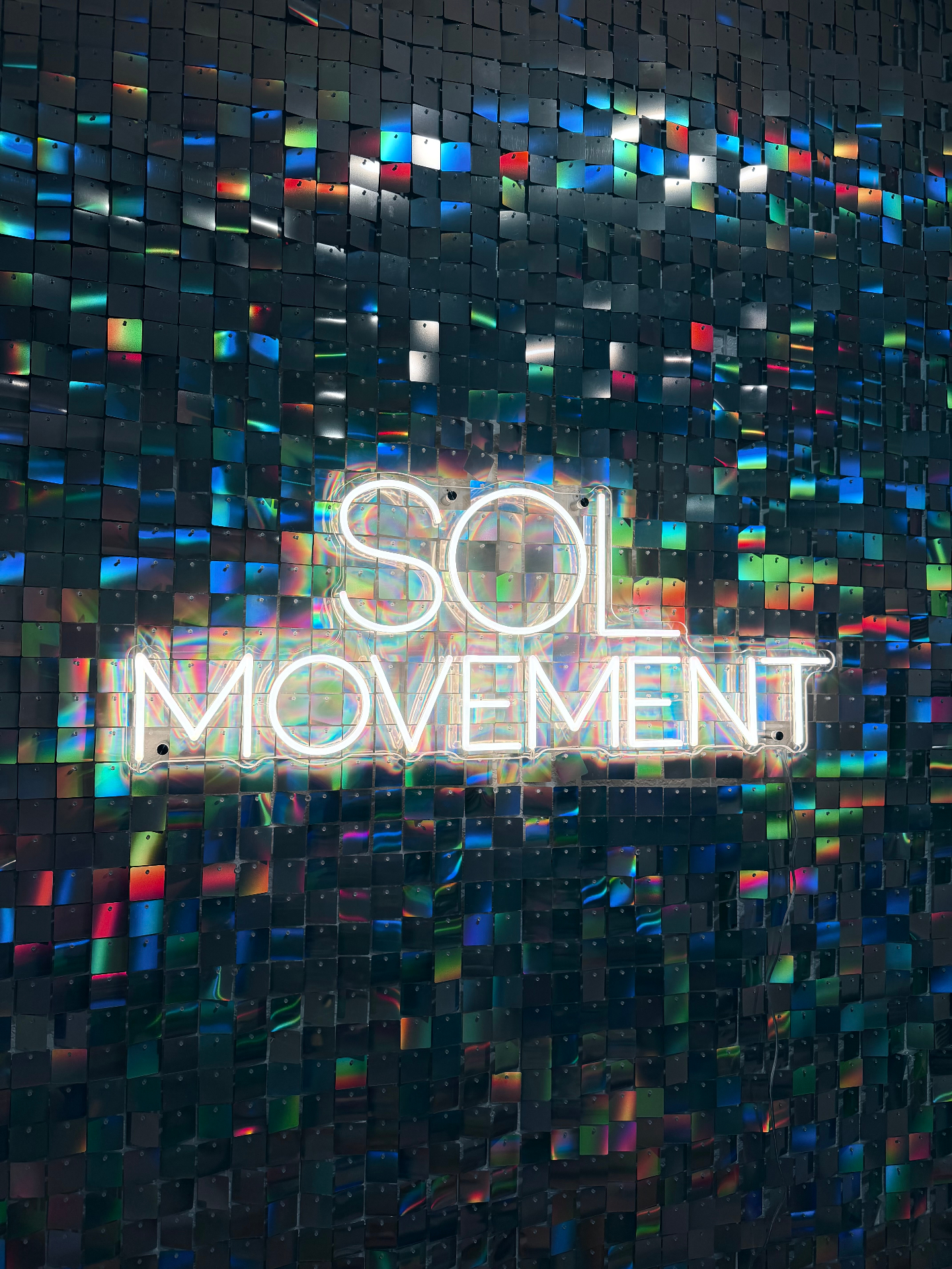 SOL Movement neon sign glowing against a shimmering iridescent sequin wall